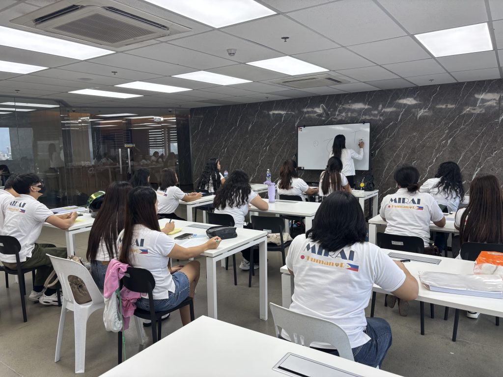 MIRAI日本語学校マカティ校の教室で、MIRAIロゴ入りの白いTシャツを着た学生たちが着席し、ホワイトボードの前に立つ講師の授業を受けている様子